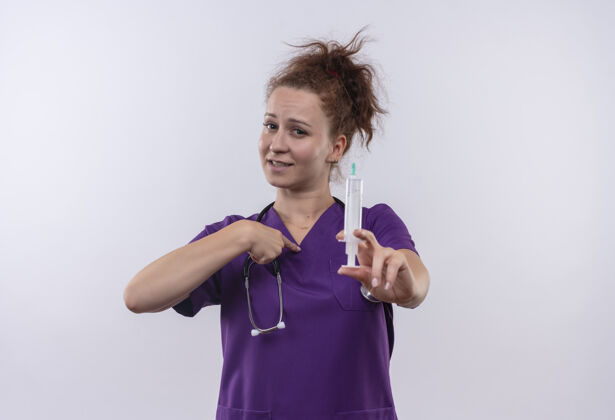 年轻人年轻的女医生身穿医疗制服 手持听诊器 手持注射器 微笑着用手指指着站在白墙上的自己注射器站着戴着