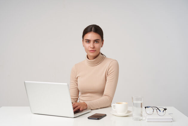 优雅迷人的年轻黑发女商人在现代办公室里工作 手里拿着笔记本电脑 双手放在键盘上 积极地看着摄像机 隔着白墙Poloneck正式玻璃