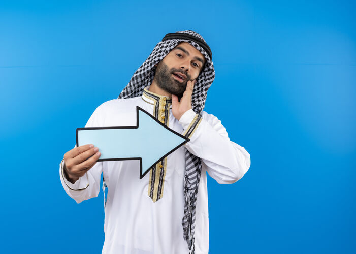 微笑身着传统服装的阿拉伯男子手持蓝色大箭头 微笑着站在蓝色的墙上指着右边箭拿着右