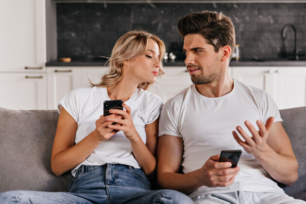 年轻卷曲的女人看着丈夫的手机迷人的白人女孩坐在沙发上拿着智能手机放松笑室内