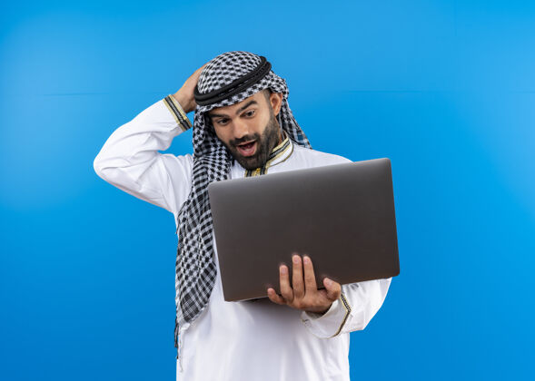 惊喜身着传统服饰的阿拉伯商人拿着笔记本电脑看着它惊讶而快乐地站在蓝色的墙上阿拉伯语站持有