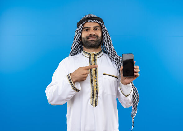 手机一位身着传统服装的阿拉伯男子站在蓝色的墙上 用手指着智能手机 自信地微笑着穿着站立指点