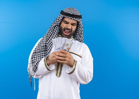 微笑身着传统服装的阿拉伯男子手持现金 站在蓝色的墙壁上愉快地微笑着站着男人欢呼