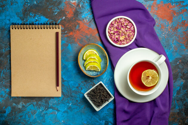 桌布俯视图一杯茶碗与干花瓣和茶片柠檬紫色桌布apencil笔记本上的蓝红色表面茶笔记本花瓣