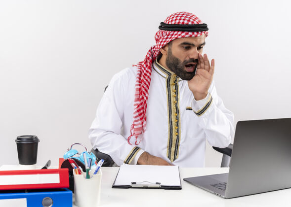 近身着传统服装的阿拉伯商人坐在桌旁 用手提电脑边喊边用手靠近嘴巴在办公室工作阿拉伯语工作穿