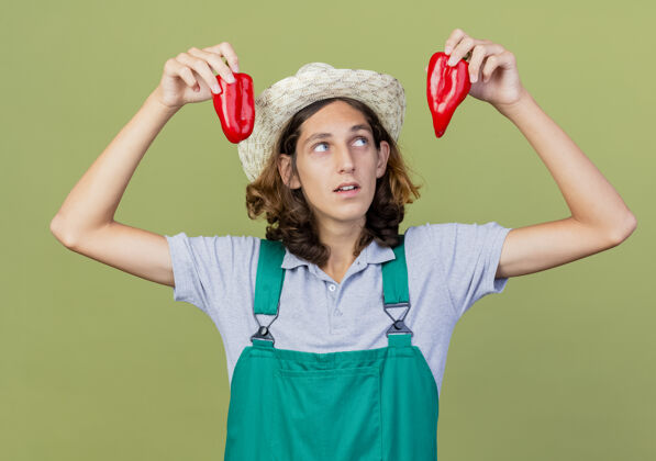连身衣年轻的园丁 穿着连体衣 戴着帽子 手里拿着红甜椒站着花园胡椒
