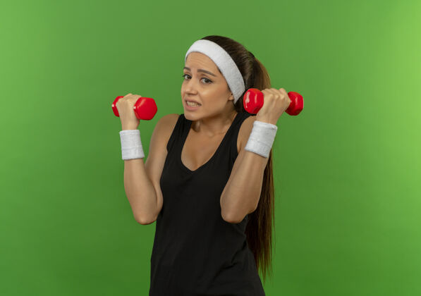 站穿着运动服的年轻健身女孩 头上带着两个哑铃 站在绿色的墙壁上做着看起来很困惑的练习运动抱头带