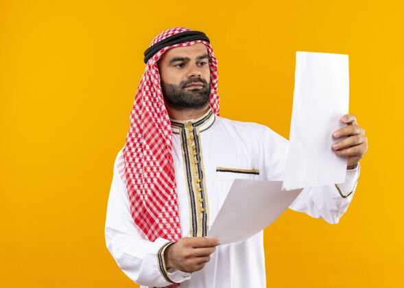 站身着传统服装的阿拉伯商人站在橙色的墙上 严肃地看着手中的文件商人阿拉伯语脸