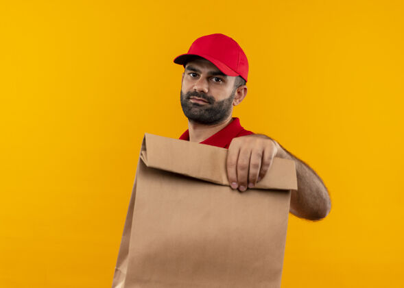 胡子一个留着胡子的送货员 穿着红色制服 戴着帽子 手里拿着纸包 严肃的脸站在橙色的墙上帽子包装拿着