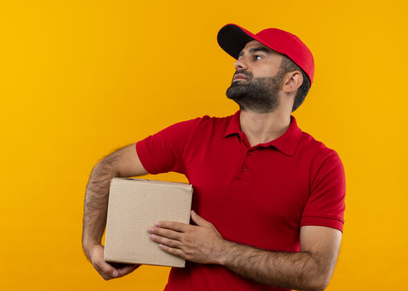 包装一个留着胡须的送货员穿着红色制服 拿着一个装帽子的盒子 站在橙色的墙上 严肃地看着旁边帽子脸送货