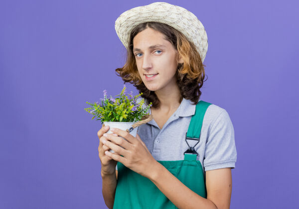 脸年轻的园丁穿着连体衣戴着帽子拿着盆栽植物微笑帽子站着