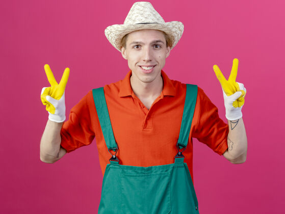橡胶年轻的园丁穿着连体衣 戴着橡胶手套的帽子 微笑着露出v字标志花园连身衣表演