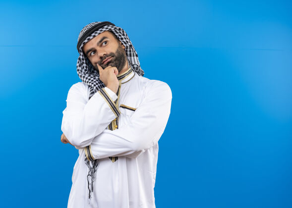 穿穿着传统服装的阿拉伯男人手放在下巴上 站在蓝色的墙上思考传统靠边阿拉伯语