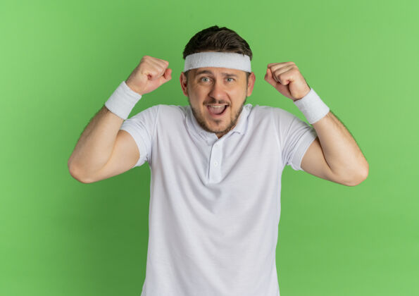 站立身穿白衬衫 头箍高举拳头的年轻健身男子站在绿色的墙壁上 快乐而兴奋运动兴奋衬衫
