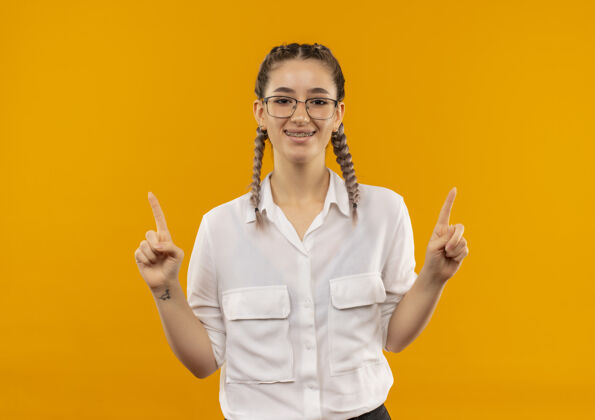 年轻人戴着眼镜 梳着辫子 穿着白衬衫的年轻女学生面带微笑地指着前面 食指竖起站在橙色的墙上眼镜女孩目录