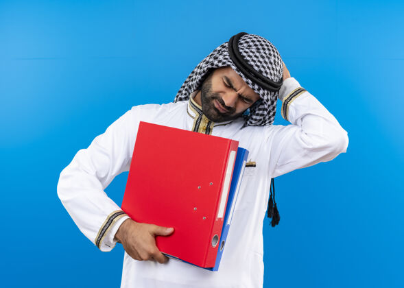 拿着身着传统服装的阿拉伯商人手持两个文件夹 站在蓝色的墙上 神情迷茫 非常焦虑阿拉伯语困惑传统