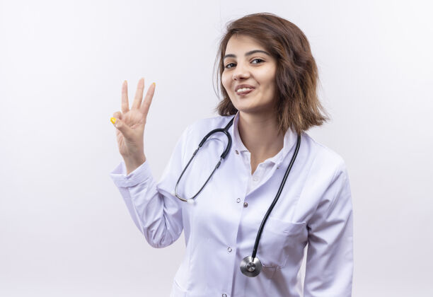 听诊器身穿白大褂 手持听诊器的年轻女医生站在白墙上微笑着指着三号手指表演医生女人