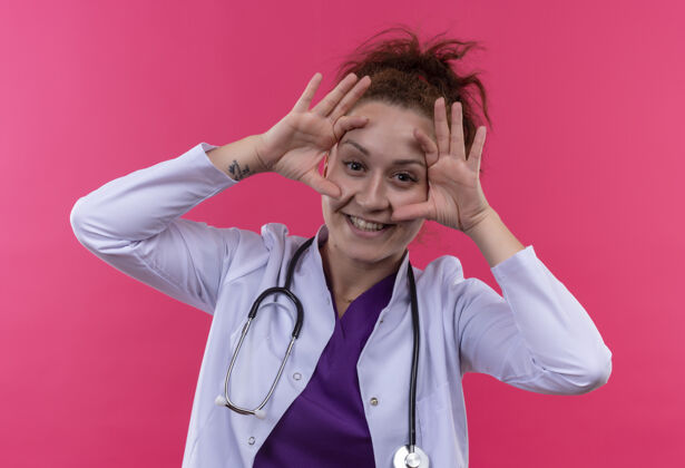 外套年轻的女医生穿着白大褂 手持听诊器睁开眼睛 手指惊讶而高兴地站在粉红色的墙上手指医生开放式