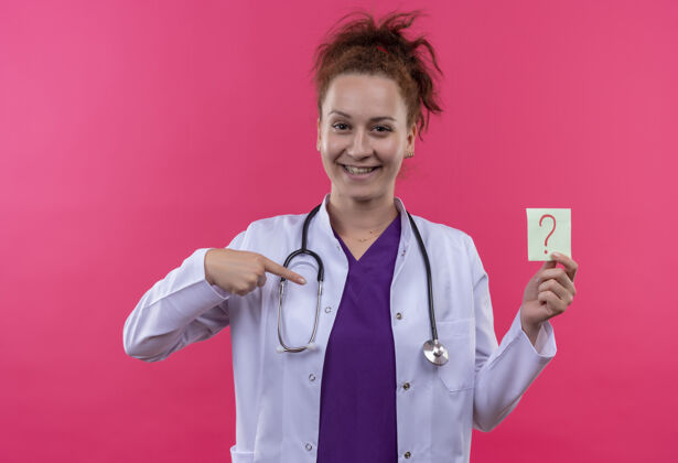 欢呼年轻的女医生穿着白大褂 手持听诊器 手拿问号纸 手指微笑着站在粉红色的墙上外套女人医生