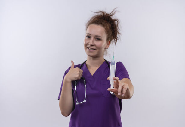 站立年轻的女医生身穿医疗制服 手持听诊器 微笑着指着注射器站在白色的墙上医生制服微笑