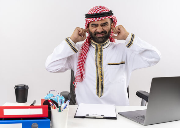 表情身着传统服装的阿拉伯商人坐在桌旁 手持笔记本电脑 用手指捂住耳朵 表情烦躁地在办公室工作工作商务人士传统