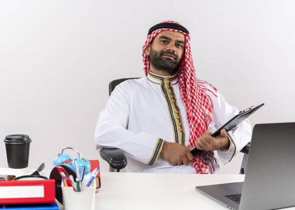 商务人士身着传统服装的阿拉伯商人坐在桌旁 手提电脑拿着剪贴板 看起来自信地在办公室工作笔记本电脑坐着电脑