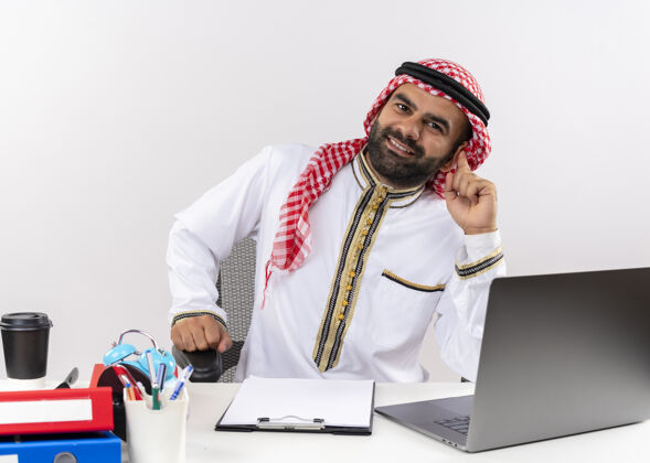 传统身着传统服装的阿拉伯商人坐在桌旁 面带笑容的手提电脑在办公室工作脸坐着桌子