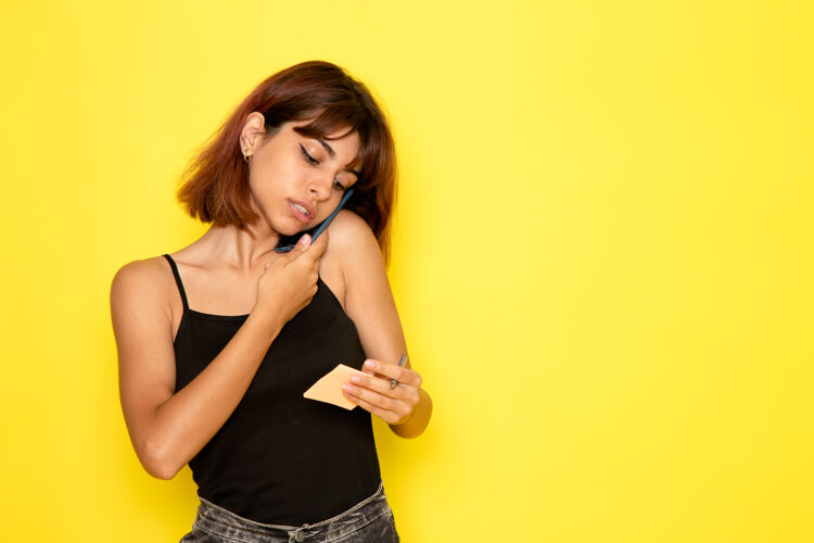 情绪黄色墙壁上穿着黑色衬衫和灰色牛仔裤的年轻女性在讲电话的前视图姿势女孩女性