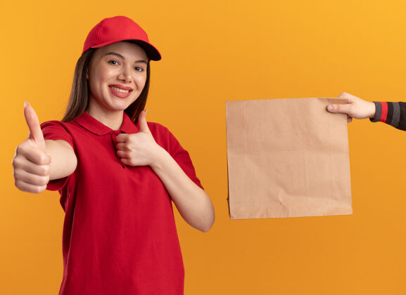 某人有人把纸包给了身着制服 面带微笑的漂亮送货员 她竖起大拇指 两只手分别放在橙色的墙上 留有复印空间漂亮包装微笑