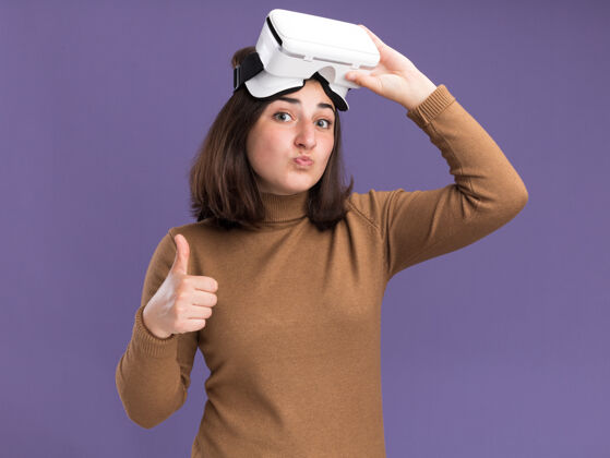 虚拟现实印象深刻的年轻漂亮的白人女孩贝雷帽举行虚拟现实耳机和竖起大拇指孤立在紫色墙上复制空间漂亮拇指印象