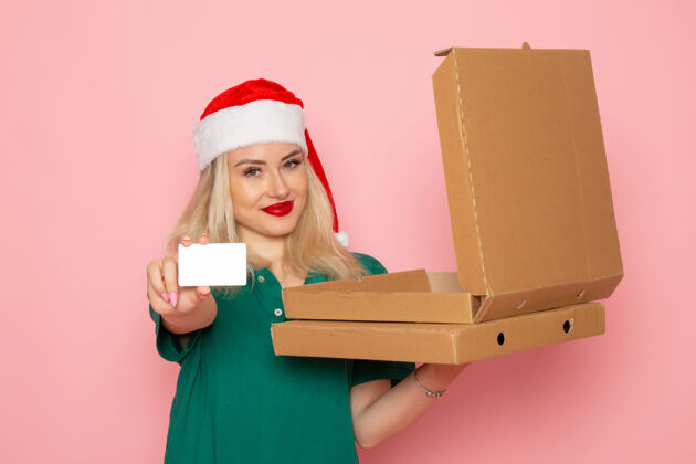 圣诞节正面图年轻女性手持银行卡和披萨盒 浅粉色墙面 彩色节日圣诞照片 工作服盒子颜色披萨