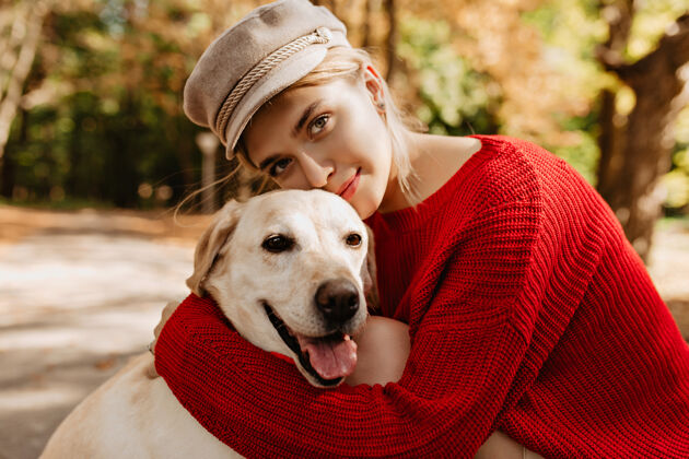 女人可爱的小女孩穿着时髦的红色套头衫 在森林里拥抱着拉布拉多犬戴着浅色帽子的金发美女和她的狗坐在公园里友谊步行者年轻