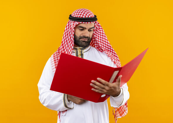 表达身着传统服装的阿拉伯商人手持打开的文件夹 严肃地看着站在橙色墙上的ot文件夹开放持有