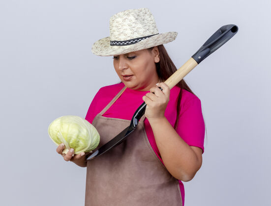 年龄围裙戴帽子的中年园丁 手里拿着卷心菜和铲子 站在白色的背景下 看上去很困惑卷心菜站着女人