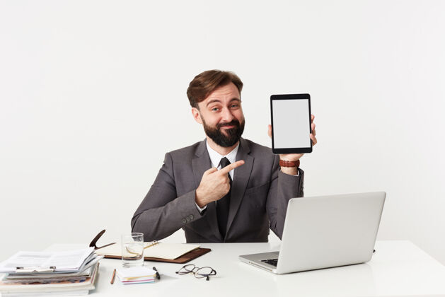 衬衫室内拍摄的是一个身穿正式服装的黑发年轻人坐在工作台前 面带讽刺的表情 手里拿着平板电脑 举手指着电脑穿着商务人士发型