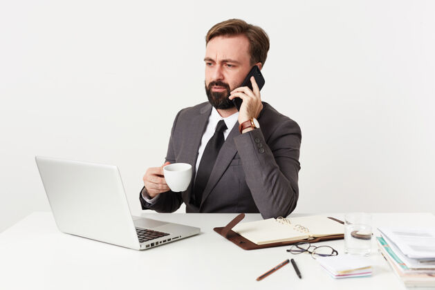 坐着严肃的留着胡子的黑发男人 穿着正式的衣服 一边打电话一边举起白杯子 一边皱眉一边看着笔记本电脑的屏幕男士笔记本困惑