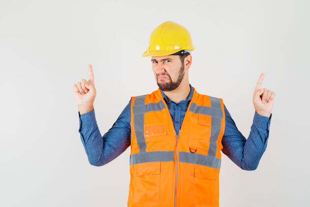 车间年轻的建筑工人指着手指在衬衫 背心 头盔和看起来阴郁 前视图工程师帽子工具
