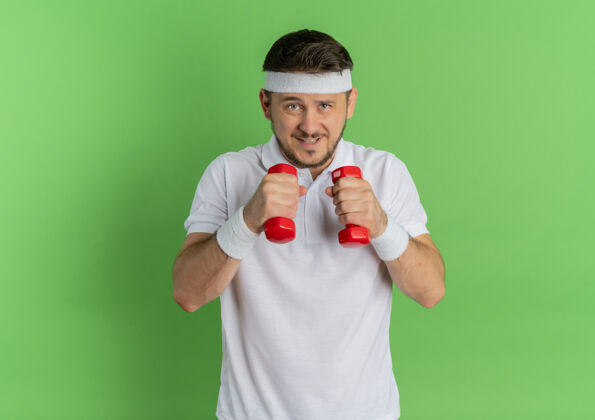 年轻人身穿白衬衫 头箍戴着哑铃的年轻健身男子站在绿色的墙上锻炼身体工作人健身
