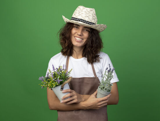 花微笑着的年轻女园丁穿着制服 戴着园艺帽 在隔离在绿墙上的花盆里拿着和交叉的花微笑花盆年轻