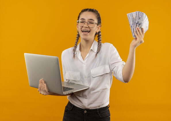 年轻戴着眼镜 梳着辫子 穿着白衬衫 拿着笔记本电脑和现金的年轻女学生站在橘色的墙上 高兴而兴奋地微笑着衬衫抱着快乐