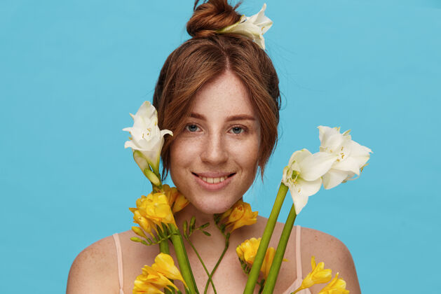 欢呼室内照片：快乐的年轻红发女性 发髻 微笑着看着相机 站在蓝色背景下 手里拿着一束鲜花表情发髻白色