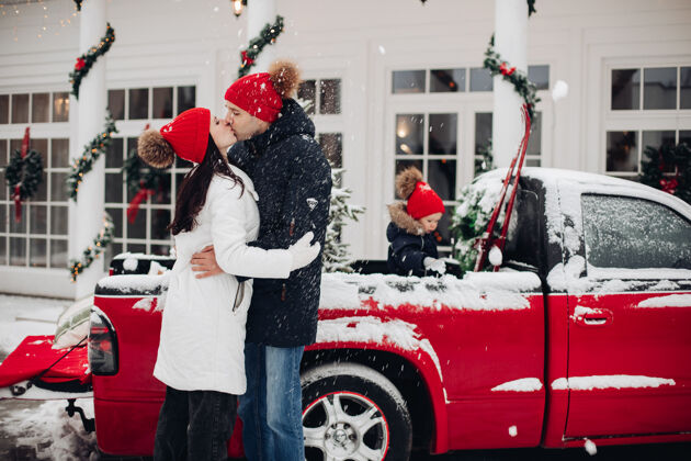 父母在户外降雪下亲吻戴着红帽子的父母汽车穿着车辆