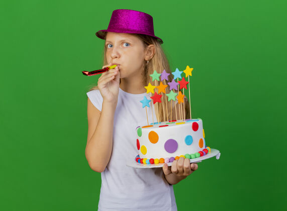 派对戴着节日帽子的漂亮小女孩拿着生日蛋糕吹口哨女孩蛋糕吹