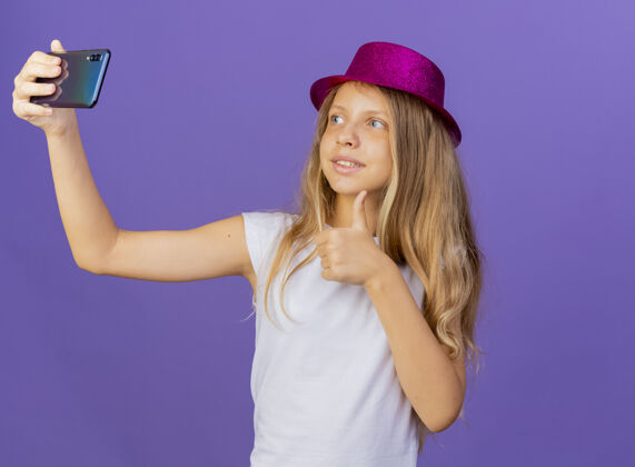 漂亮戴着节日礼帽的漂亮小女孩用智能手机自拍微笑着竖起大拇指 紫色背景下站着生日派对的概念紫色女孩小