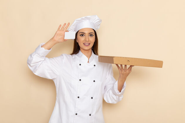 女身穿白色厨师服的年轻女厨师的正视图 手持卡片和白墙上的食物盒人年轻举行