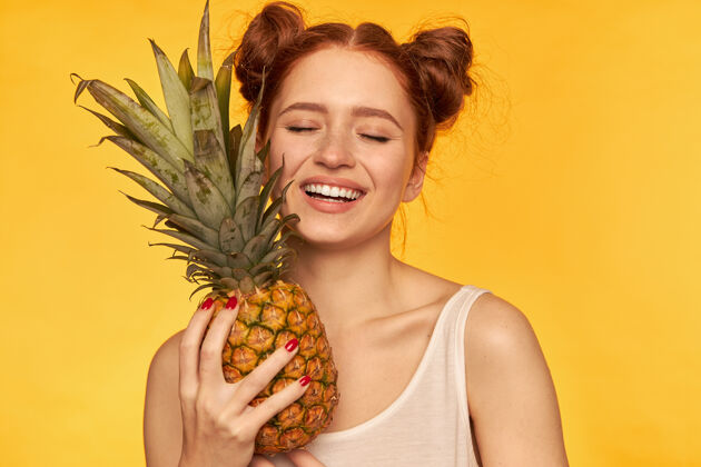休闲微笑着闭着眼睛的少女 满脸幸福的红发女人 带着两个发髻 穿着白衬衫 手里拿着菠萝 健康的生活方式 孤立地站在黄色的墙上面部长相闭眼