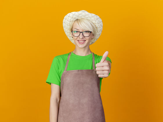 女人围裙上戴着帽子 留着短发的年轻女园丁站在橙色的背景下 面带微笑 竖起大拇指看着镜头橙色围裙帽子