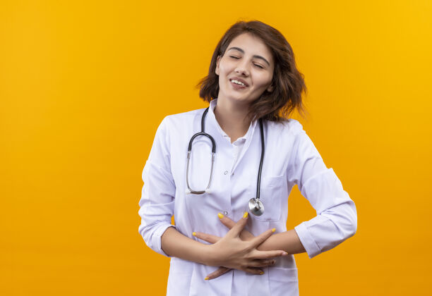 腹部一位年轻的女医生 身穿白大褂 戴着听诊器 闭着眼睛 微笑着站在橙色的墙上 摸着她的肚子封闭外套女人