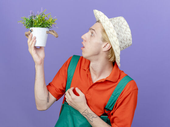 抱着年轻的园丁穿着连体衣戴着帽子拿着盆栽植物惊讶地看着它植物站着男人