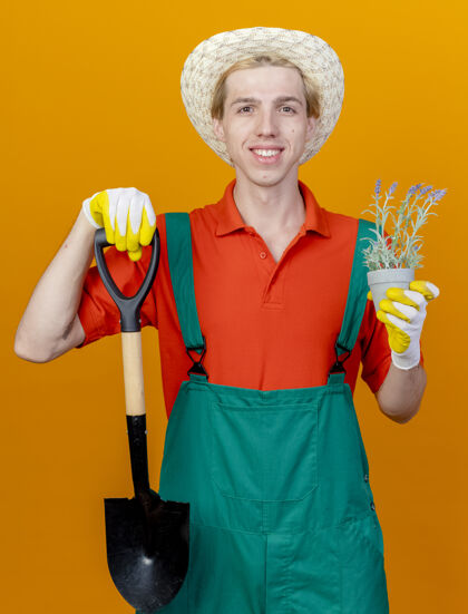 锅年轻的园丁穿着连体衣戴着帽子拿着铲子和盆栽植物穿帽子微笑